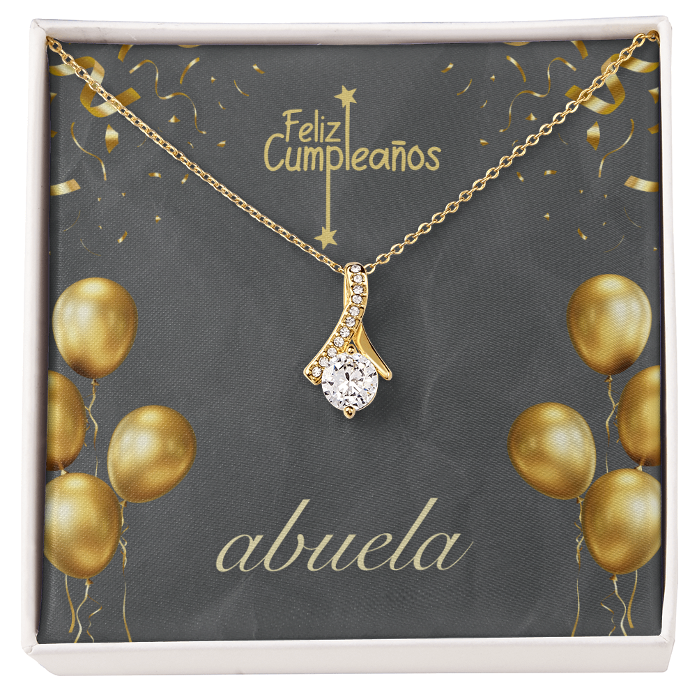 Una cadena de oro con un dije de cristal brillante en forma de gota, presentada en una tarjeta de regalo temática de fondo gris oscuro. La tarjeta muestra el texto en español "Feliz Cumpleaños" en la parte superior y "abuela" en letra cursiva blanca en la parte inferior, decorada con ilustraciones de globos y confeti dorados.