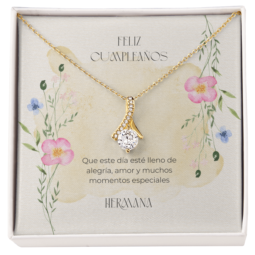 Collar dorado con un dije en forma de cinta decorado con cristales y una gran piedra brillante central, presentado en una caja blanca sobre una tarjeta con ilustraciones florales en acuarela y un mensaje de cumpleaños en español: "Feliz Cumpleaños. Que este día esté lleno de alegría, amor y muchos momentos especiales. Hermana."