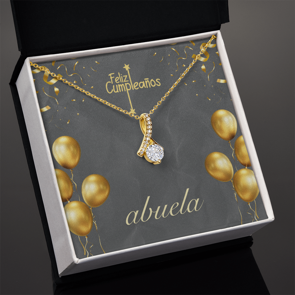 Una cadena de oro con un dije de cristal brillante en forma de gota, presentada en una caja de regalo premium de color negro con interior blanco. La tarjeta de fondo es de color gris oscuro y muestra el texto en español "Feliz Cumpleaños" en la parte superior y "abuela" en letra cursiva blanca en la parte inferior, decorada con ilustraciones de globos y confeti dorados.