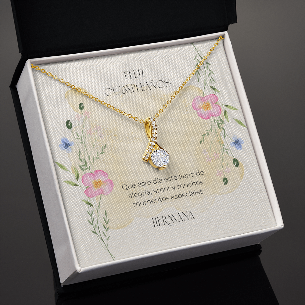 Vista en ángulo de un collar dorado con un dije en forma de cinta entrelazada y piedra brillante central, presentado en una caja de regalo negra abierta. El fondo interior muestra una tarjeta con flores silvestres y la dedicatoria en español: "Feliz Cumpleaños. Que este día esté lleno de alegría, amor y muchos momentos especiales. Hermana." 