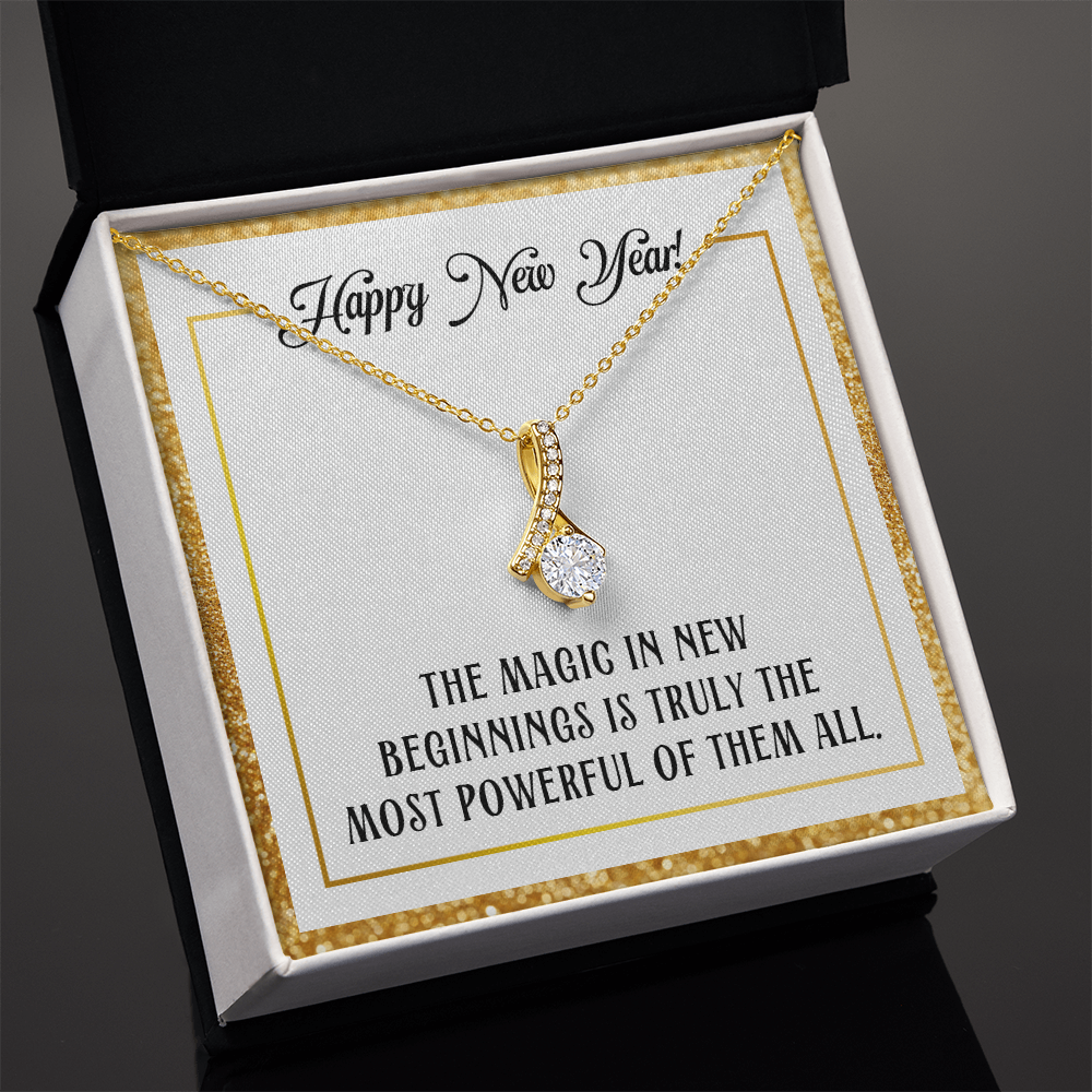 A gold-colored pendant necklace featuring a large, round-cut central crystal dangling from a ribbon-shaped bail encrusted with smaller crystals. The necklace is displayed inside a square white gift box with a sparkling gold glitter border. The necklace rests on a white card that reads "Happy New Year!" at the top in cursive and "THE MAGIC IN NEW BEGINNINGS IS TRULY THE MOST POWERFUL OF THEM ALL." at the bottom in bold, uppercase block lettering.

