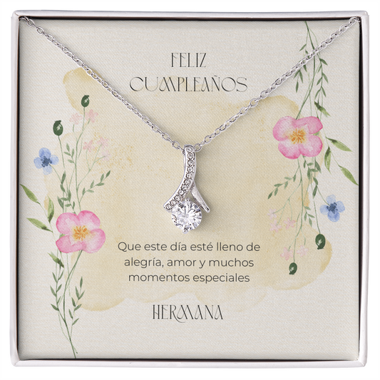 Collar con colgante de plata en forma de lazo y una gran circonita cúbica redonda, presentado sobre una tarjeta de regalo decorativa con motivos florales dentro de una caja blanca. La tarjeta, de color crema, está ilustrada con delicadas flores silvestres pintadas en acuarela (en tonos rosa y azul) y lleva un mensaje de cumpleaños en español: "Feliz Cumpleaños. Que este día esté lleno de alegría, amor y muchos momentos especiales. Hermana".