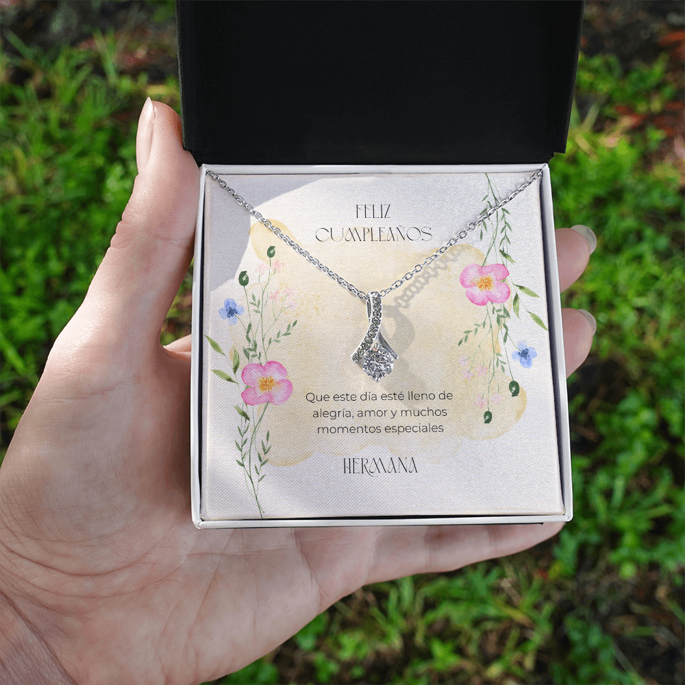 Persona sosteniendo en la mano una caja de regalo negra abierta con un collar plateado en su interior, con un fondo exterior difuminado de césped verde. El dije en forma de cinta brillante reposa sobre una tarjeta decorada con flores silvestres en acuarela y el mensaje en español: "Feliz Cumpleaños. Que este día esté lleno de alegría, amor y muchos momentos especiales. Hermana." 