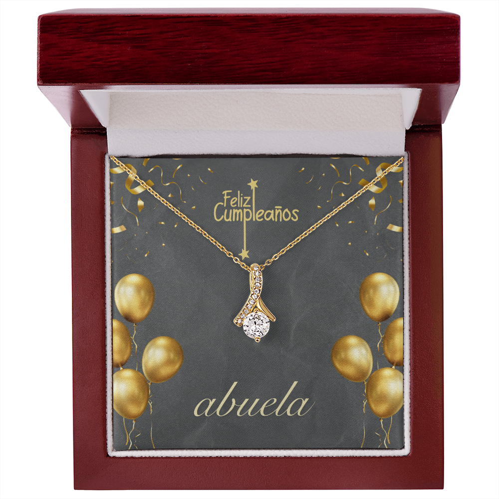 Una cadena de oro con un dije de cristal brillante en forma de gota, presentada dentro de una elegante caja de regalo de madera pulida color caoba con interior blanco. La tarjeta de fondo es de color gris oscuro y muestra el texto en español "Feliz Cumpleaños" en la parte superior y "abuela" en letra cursiva blanca en la parte inferior, decorada con ilustraciones de globos y confeti dorados.