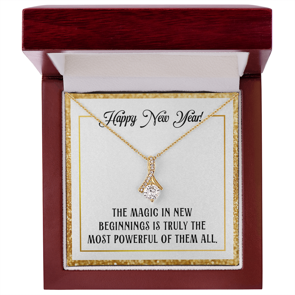 A gold-colored pendant necklace featuring a large, round-cut central crystal dangling from a ribbon-shaped bail encrusted with smaller crystals. The necklace is displayed inside a square dark red wood gift box with a sparkling gold glitter border. The necklace rests on a white card that reads "Happy New Year!" at the top in cursive and "THE MAGIC IN NEW BEGINNINGS IS TRULY THE MOST POWERFUL OF THEM ALL." at the bottom in bold, uppercase block lettering.
