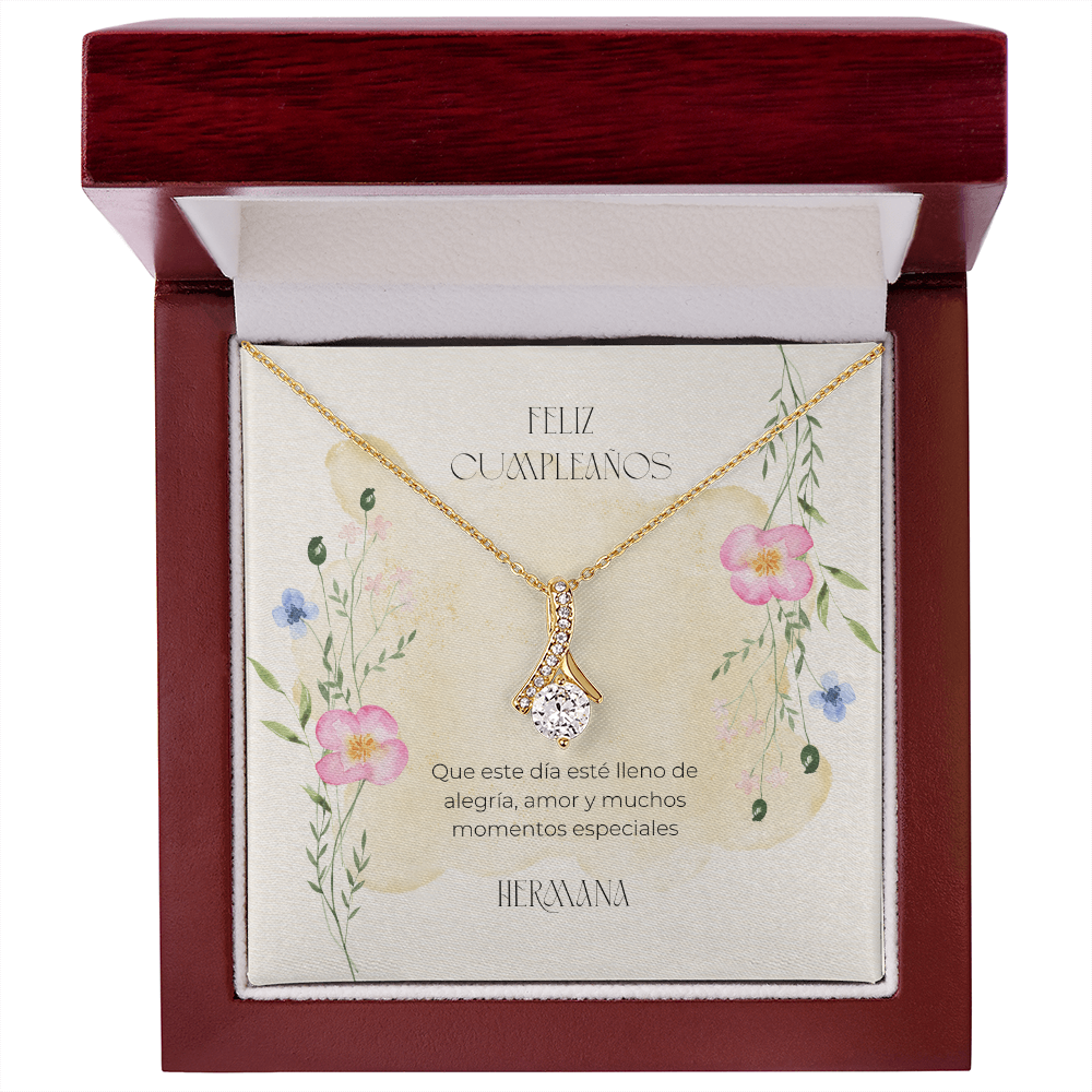 Collar dorado con dije en forma de cinta y piedra brillante central, exhibido dentro de una caja de madera de lujo abierta con acabado de caoba. La tarjeta interior con diseño de flores silvestres en acuarela incluye el mensaje en español: "Feliz Cumpleaños. Que este día esté lleno de alegría, amor y muchos momentos especiales. Hermana."