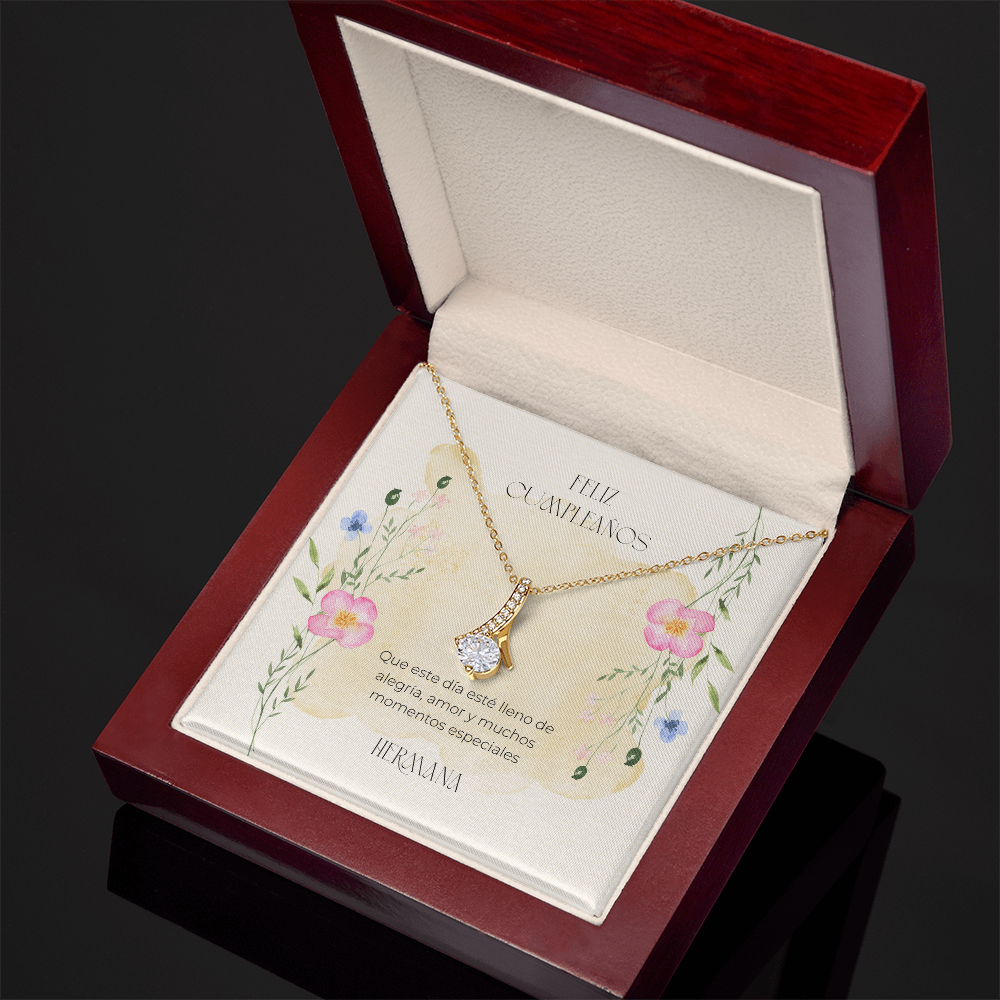 Vista en perspectiva diagonal de un collar dorado con un dije en forma de cinta entrelazada y piedra brillante, presentado en una caja de madera de lujo abierta con acabado caoba. La tarjeta interior muestra ilustraciones florales y la dedicatoria en español: "Feliz Cumpleaños. Que este día esté lleno de alegría, amor y muchos momentos especiales. Hermana."