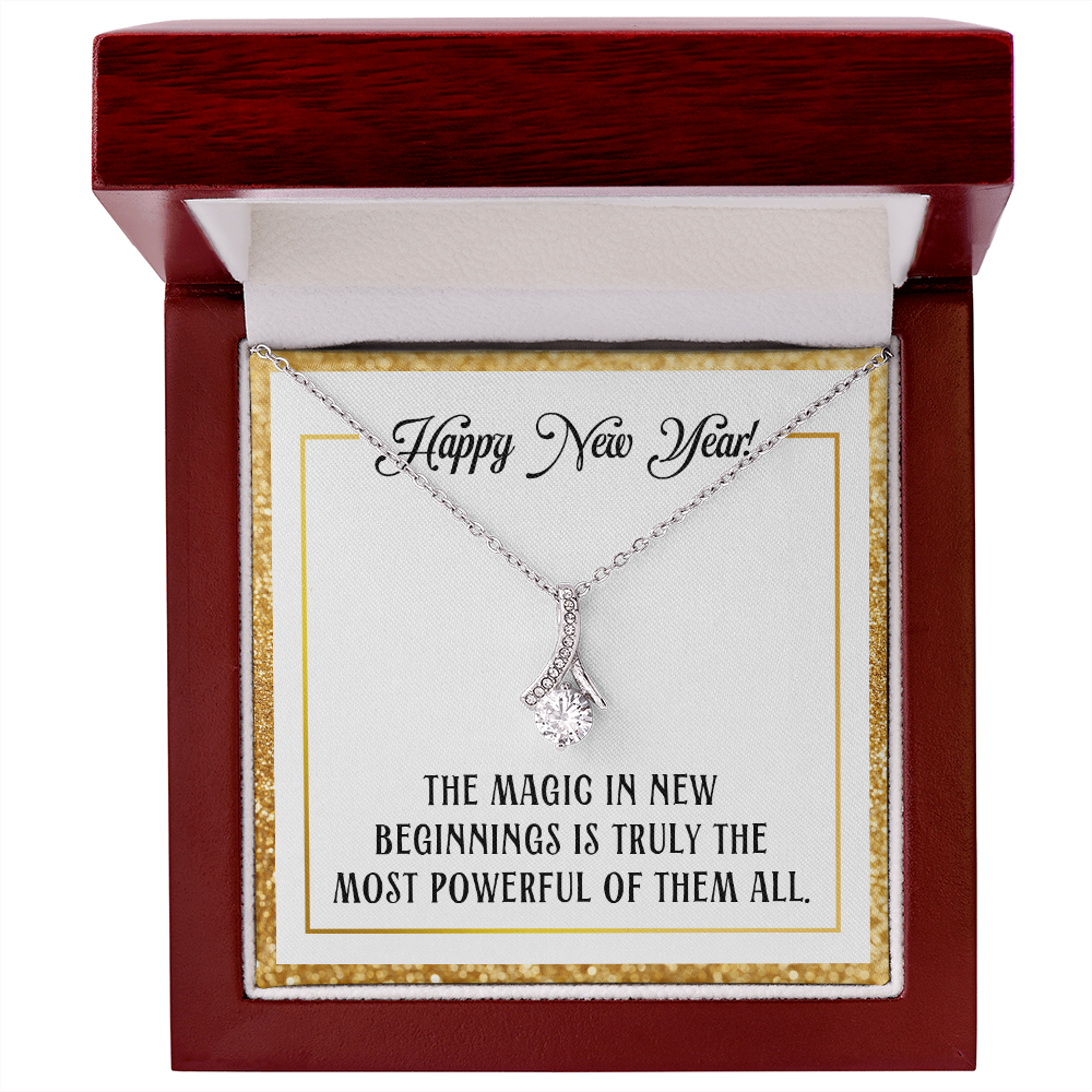 A silver-colored pendant necklace featuring a large, round-cut central crystal dangling from a ribbon-shaped bail encrusted with smaller crystals. The necklace is displayed inside a dark red wood gift box with a sparkling gold glitter border. The necklace rests on a white card that reads "Happy New Year!" at the top in cursive and "THE MAGIC IN NEW BEGINNINGS IS TRULY THE MOST POWERFUL OF THEM ALL." at the bottom in bold, uppercase block lettering.
