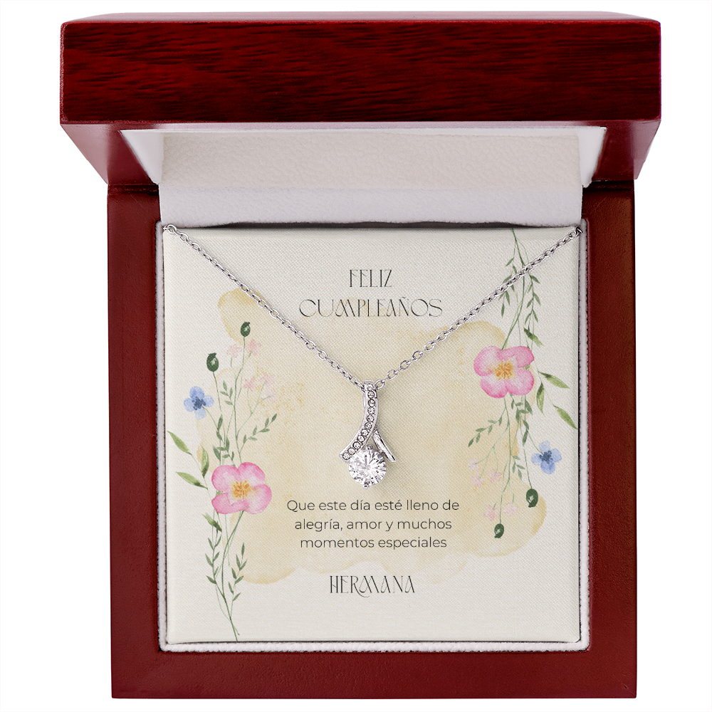 Collar de plata con dije en forma de cinta y piedra brillante central, presentado dentro de una caja de madera de lujo abierta con acabado de caoba. La tarjeta interior de fondo crema con flores silvestres muestra el mensaje en español: "Feliz Cumpleaños. Que este día esté lleno de alegría, amor y muchos momentos especiales. Hermana."