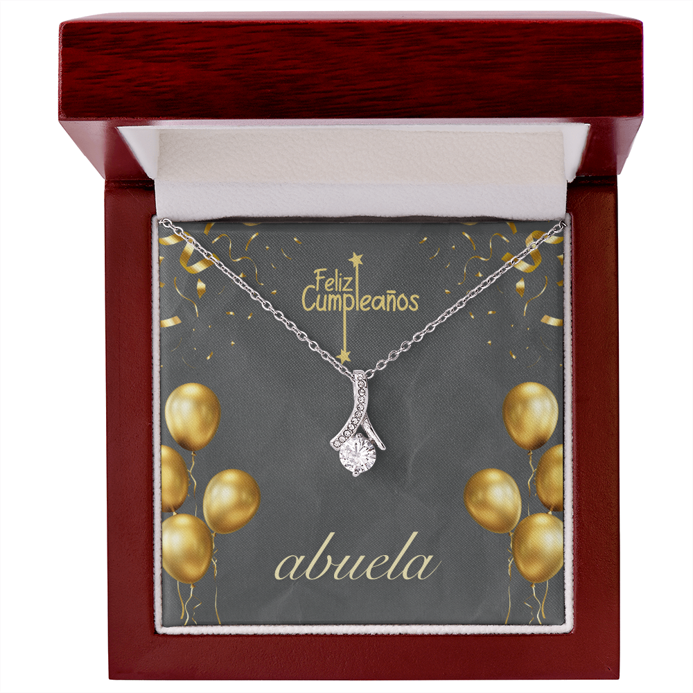Una cadena de plata con un dije de cristal brillante en forma de gota, presentada dentro de una elegante caja de regalo de madera pulida color caoba con interior blanco. La tarjeta de fondo es de color gris oscuro y muestra el texto en español "Feliz Cumpleaños" en la parte superior y "abuela" en letra cursiva blanca en la parte inferior, decorada con ilustraciones de globos y confeti dorados.