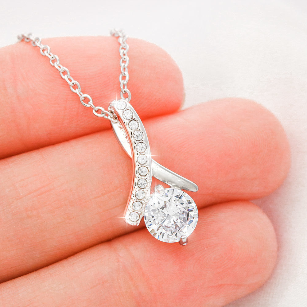 a silver toned ribbon pendant with cubic zirconia crystals on a silver chain resting on the tip of a woman's hand.