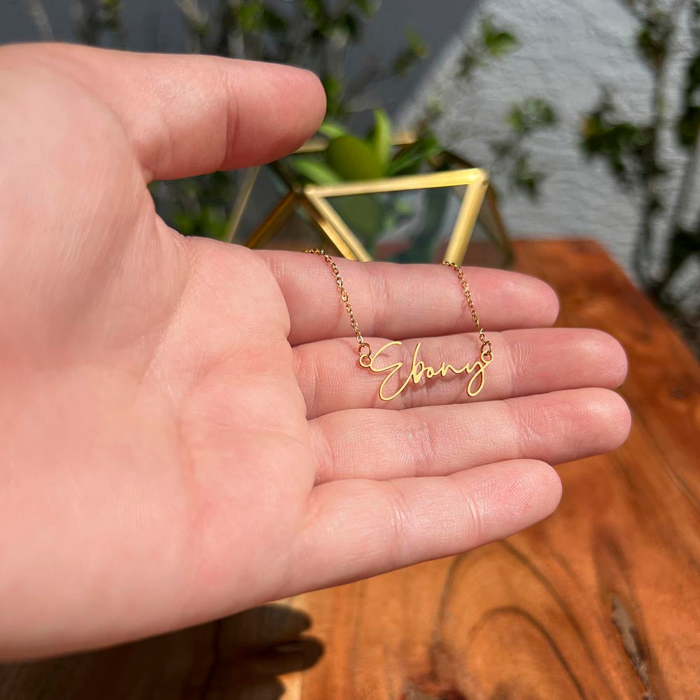 A personalized gold necklace displayed on a man's hand with a wood and green leaf background.