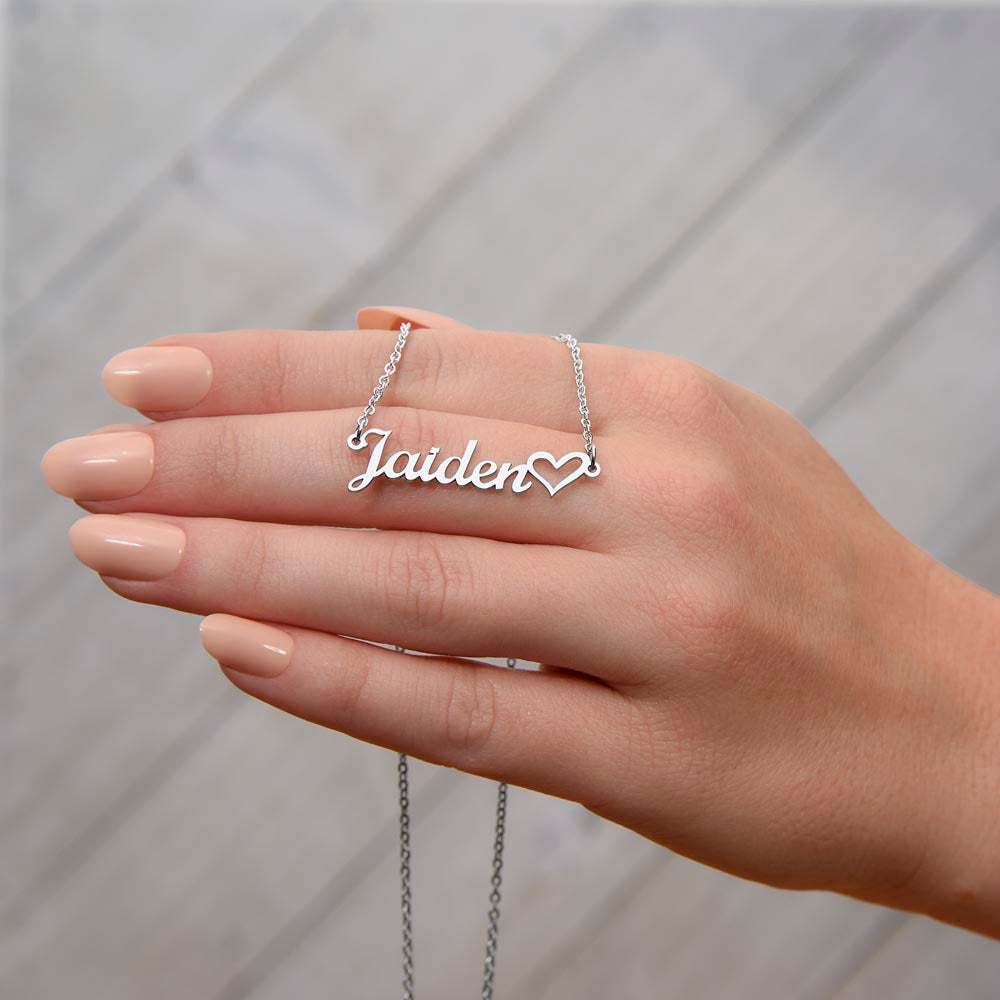 Personalized silver necklace with a heart-shaped nameplate, presented on a woman's hand with light peach nails and a light gray accent background.