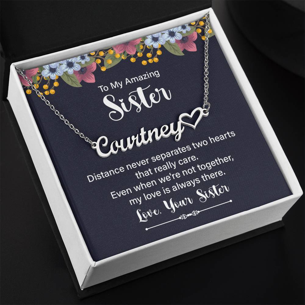 Personalized silver necklace with a heart-shaped nameplate, presented in a square gift box with a navy card featuring floral accents and a sentimental message for a sister in a white gift box with a black background.