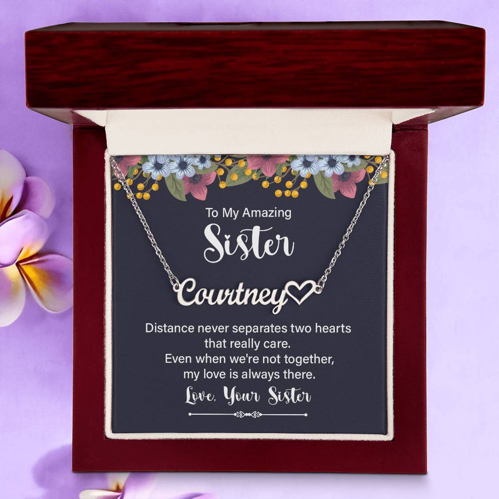 Personalized silver necklace with a heart-shaped nameplate, presented in a square gift box with a navy card featuring floral accents and a sentimental message for a sister in a dark wood gift box with a lavender floral background.
