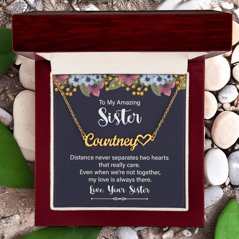 Personalized gold necklace with a heart-shaped nameplate, presented in a square gift box with a navy card featuring floral accents and a sentimental message for a sister in a dark wood gift box with a rock and green leaf background.