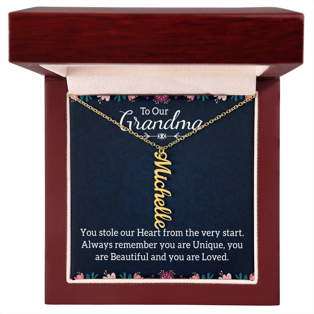 A personalized gold nameplate necklace displayed on a dark navy gift card with floral accents and a sentimental message for 'Grandma' in a dark wood gift box with a white background.