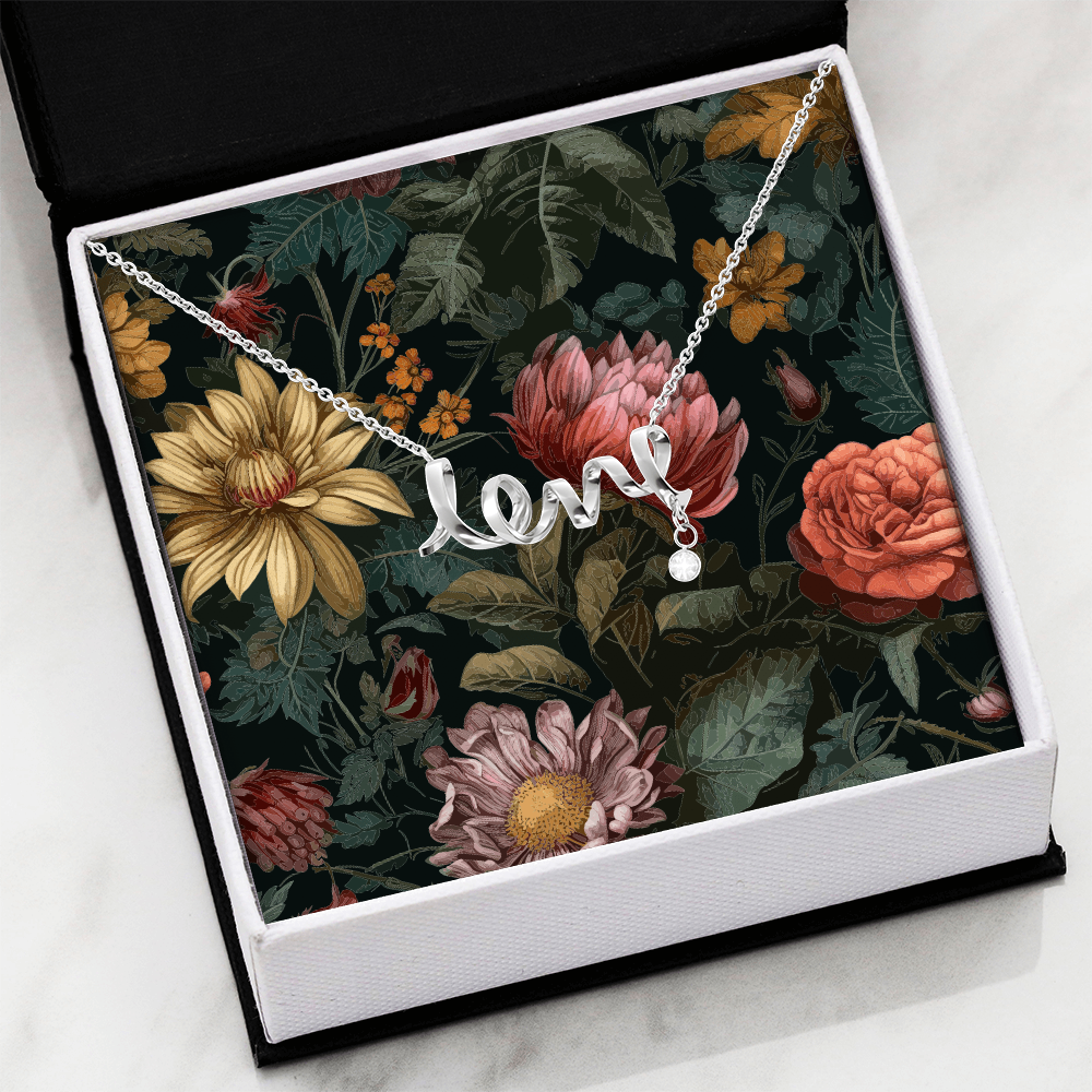 An angled, top-down view of a silver script "love" necklace with a dangling round crystal charm, presented inside an open black-trimmed white gift box against a dark, vintage-style floral pattern backdrop of yellow, pink, and orange blossoms.