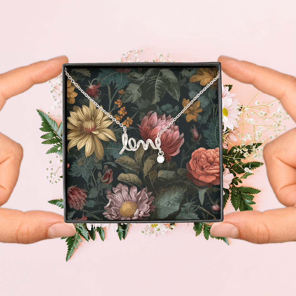 A top-down view of two hands holding the sides of an open black jewelry box displaying a silver script "love" necklace with a small dangling round crystal charm, framed by a soft bouquet of white and pink flowers on a light pink background.