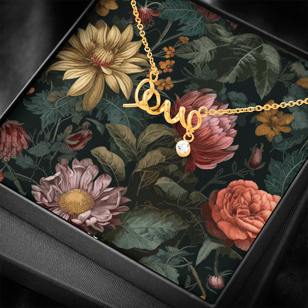 A close-up, angled view of a gold script "love" necklace with a dangling round crystal charm, presented on a dark vintage floral card inside a textured black gift box.