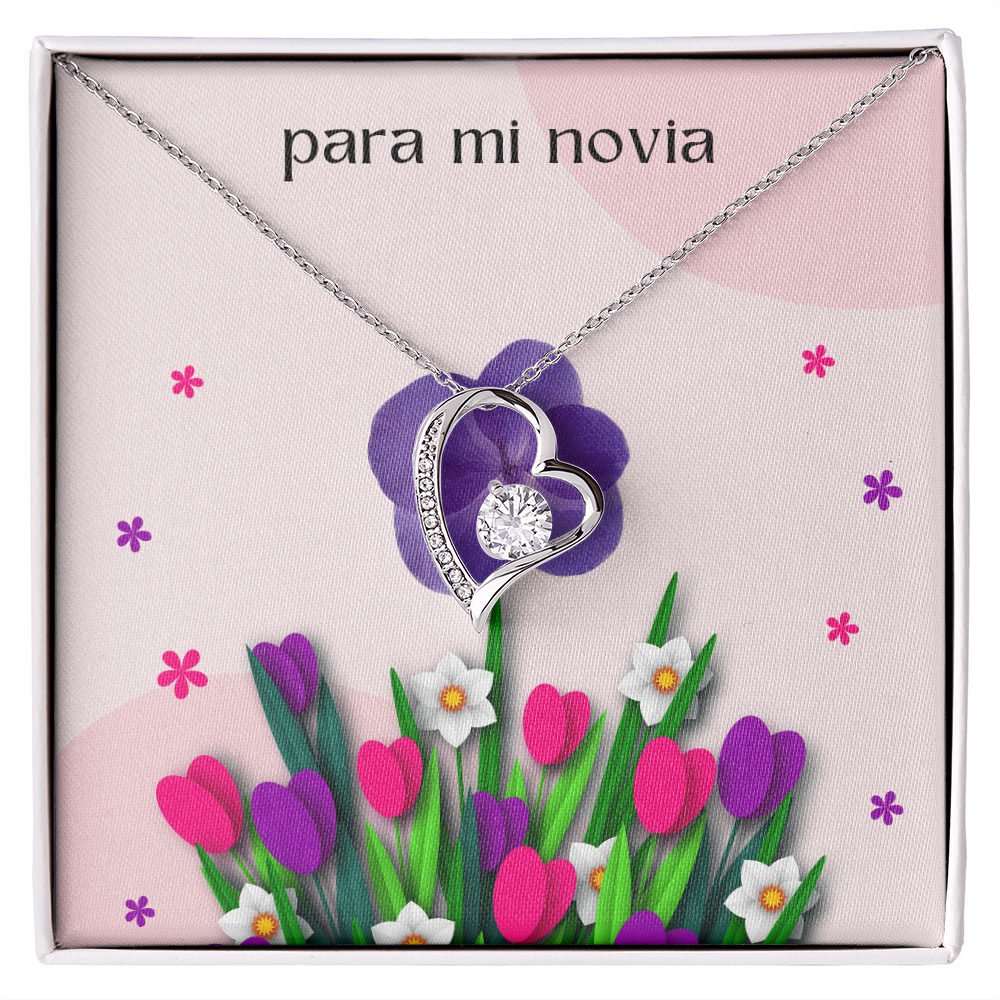 Collar plateado con dije de corazón y cristal brillante sobre una tarjeta de regalo floral rosa que dice "para mi novia" en letras minúsculas.