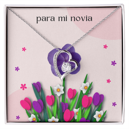 Collar plateado con dije de corazón y cristal brillante sobre una tarjeta de regalo floral rosa que dice "para mi novia" en letras minúsculas.