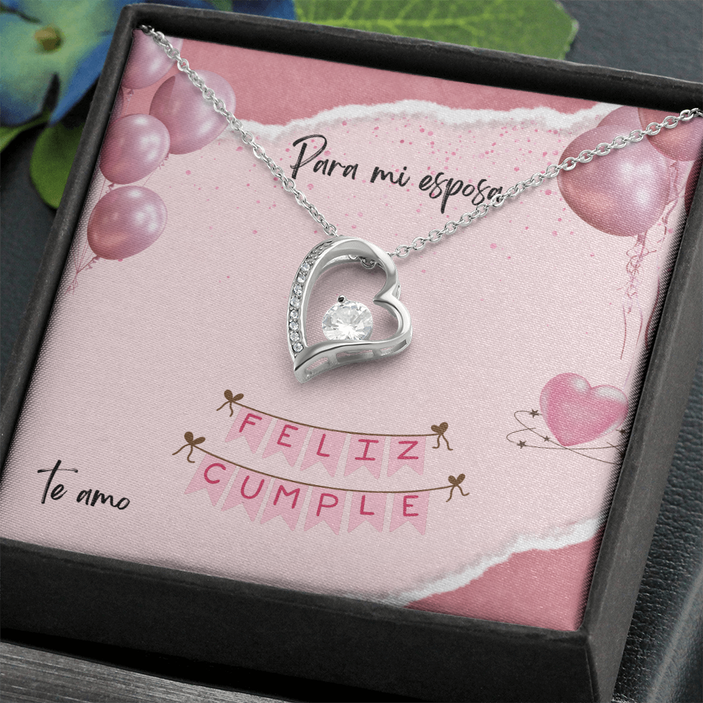Collar plateado con dije de corazón y cristal brillante, presentado en una caja de regalo oscura inclinada con una tarjeta rosa que dice "Para mi esposa, Feliz Cumple, Te amo".