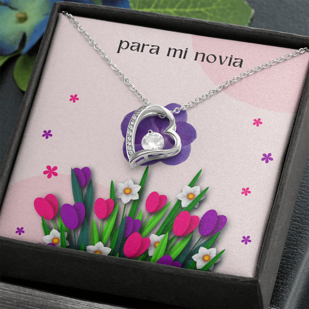 Collar plateado con dije de corazón y cristal brillante, presentado en una caja de regalo oscura e inclinada con una tarjeta floral rosa que dice "para mi novia".