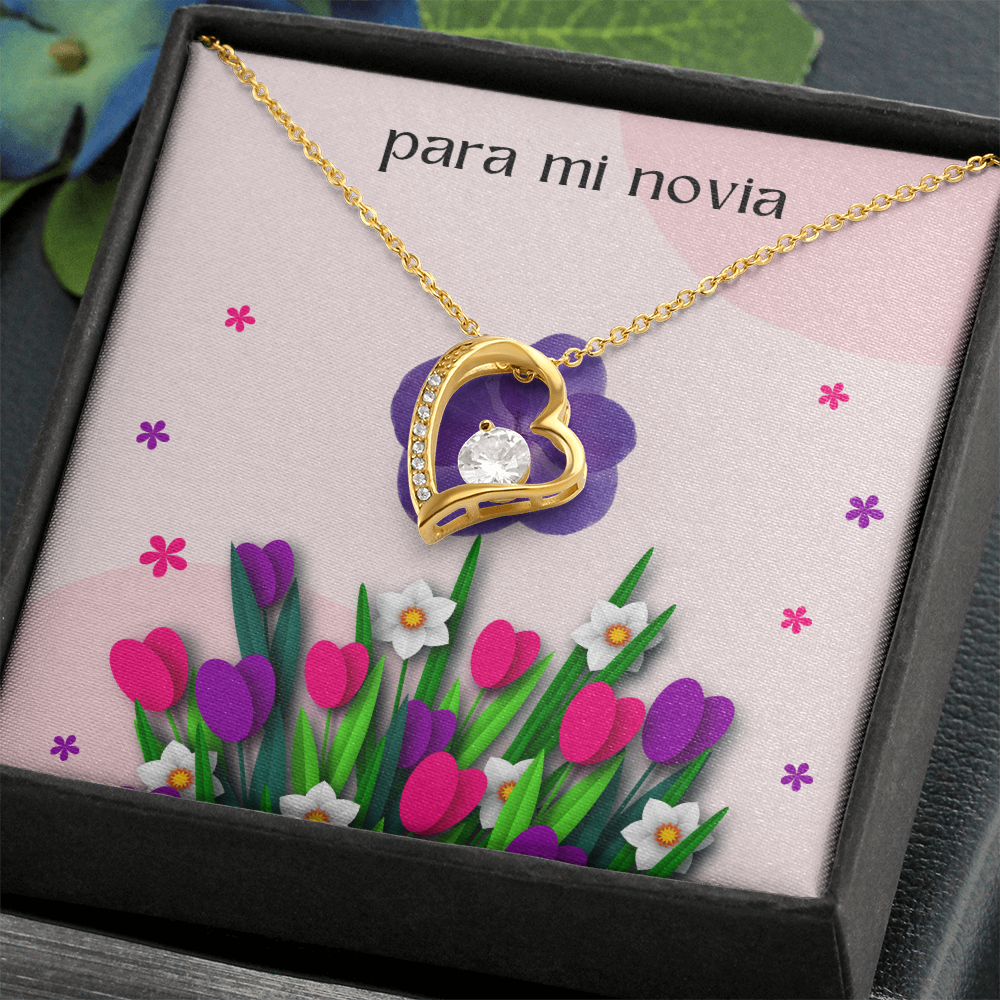 Collar de oro con dije de corazón y cristal brillante, presentado en una caja de regalo oscura e inclinada con una tarjeta floral rosa que dice "para mi novia".