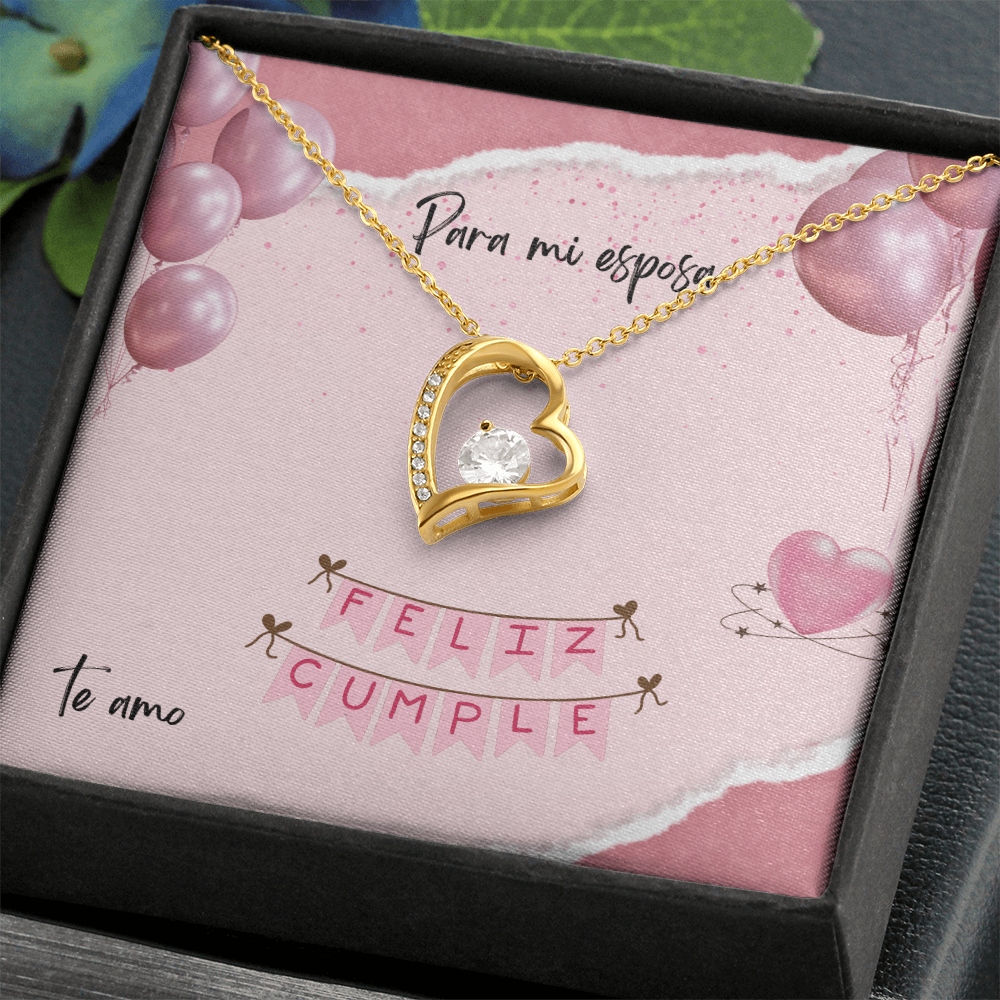 Collar de oro con un dije de corazón y un cristal brillante, presentado en una caja de regalo oscura e inclinada con una tarjeta rosa que dice "Para mi esposa, Feliz Cumple, Te amo".