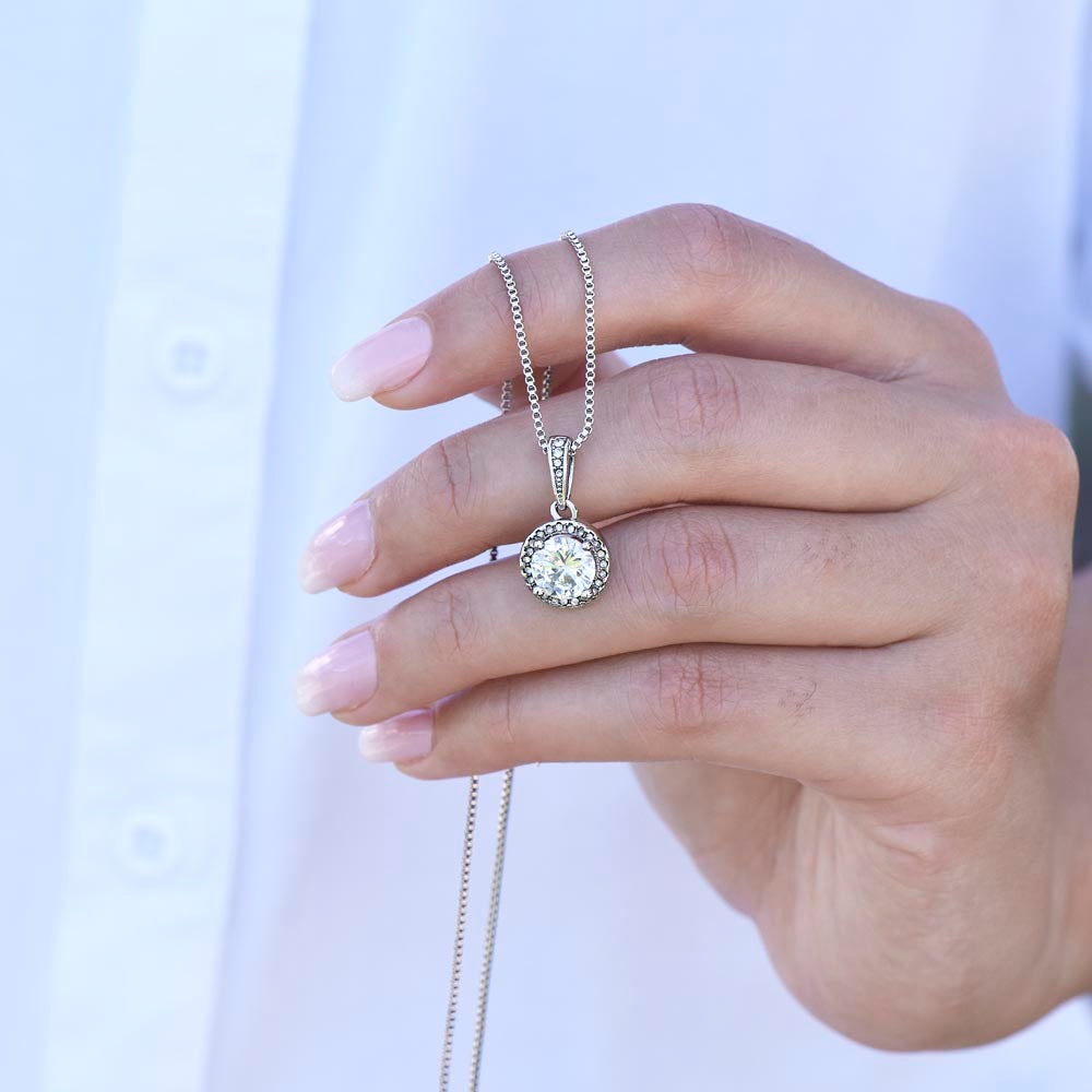 A close-up of a hand with a soft pink ombré manicure gently holding a silver round halo pendant necklace with a sparkling center stone, set against a crisp white shirt background.