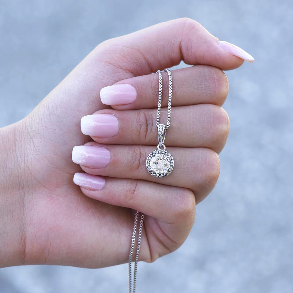 A woman's hand with white and pink ombré manicured nails holding up a silver round halo pendant necklace with a brilliant-cut center gemstone, dangling against a blurred gray background.