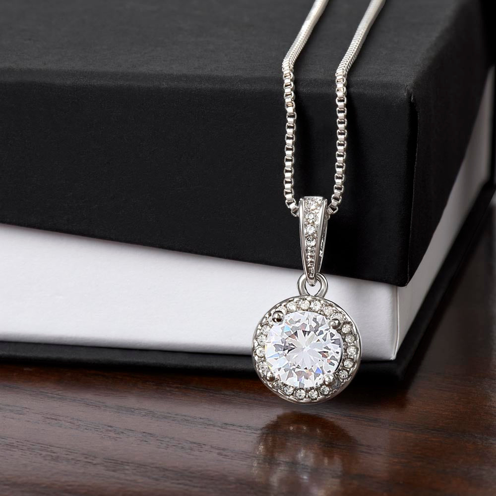 A silver round halo pendant necklace with a large, brilliant-cut center stone surrounded by a border of smaller stones, hanging on a silver box chain in front of a stacked black and white gift box.