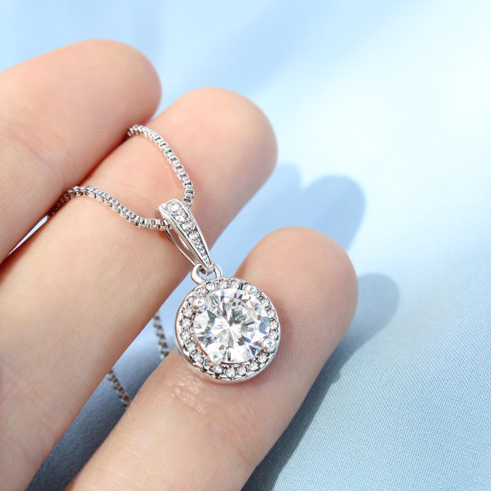 A person's hand holding a silver round halo pendant necklace with a large, sparkling central gemstone and a pavé stone border, resting against a soft light blue background.