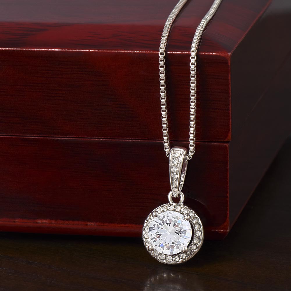 A round halo pendant necklace with a large, brilliant-cut central gemstone surrounded by smaller pavé-set stones, hanging from a delicate silver box chain against a dark reddish-brown wooden gift box.