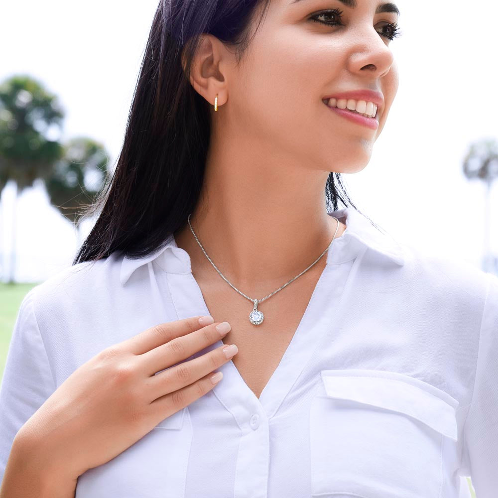 A smiling woman with dark hair wearing a white button-up shirt, showcasing a silver round halo pendant necklace and a small gold huggie earring, while looking slightly off-camera in an outdoor setting.