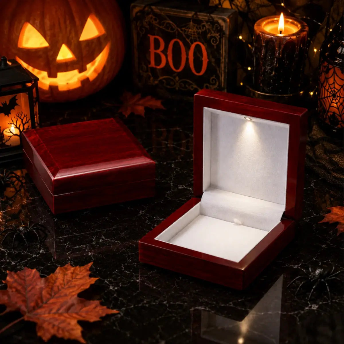A Halloween-themed product photo showing a glossy dark red wooden jewelry box, one closed and one open with a built-in warm white LED light illuminating the interior. The boxes are displayed on a dark marble surface surrounded by seasonal props including a lit jack-o'-lantern, a candle, autumn leaves, and a 'BOO' sign.