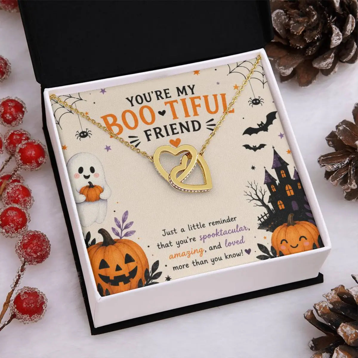 A Halloween-themed gift box containing a gold necklace with two interlocking heart pendants. The necklace is displayed on a cream-colored card decorated with illustrations of a ghost, jack-o'-lantern, haunted house, bats, and spiderwebs, with the text 'YOU'RE MY BOOTIFUL FRIEND'.