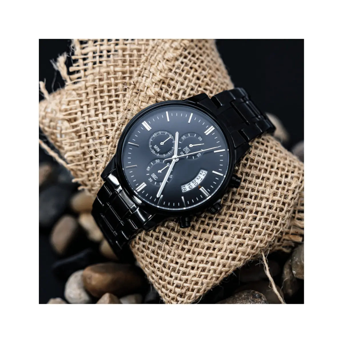 A black analog watch with a metal link bracelet and silver-tone hands, displayed on a burlap-wrapped roll with small stones around it.