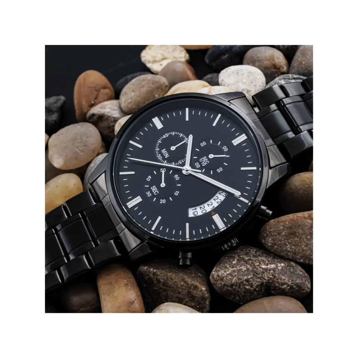 A black analog watch with a black metal link bracelet, featuring a black dial with silver-tone hands, markers, and subdials, resting on a bed of smooth river stones.