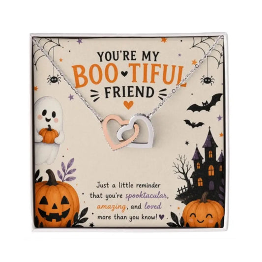 A Halloween-themed gift card featuring a silver and rose gold double heart pendant necklace with cubic zirconia. The card is decorated with spooky illustrations including a ghost, jack-o'-lanterns, a haunted house, bats, and spiders, with the text 'YOU'RE MY BOOTIFUL FRIEND'.