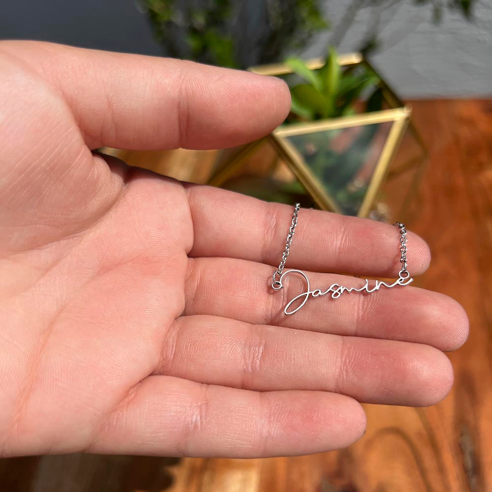 A man's hand displaying a silver personalized signature style name pendant necklace against a background of green leaves and wood.