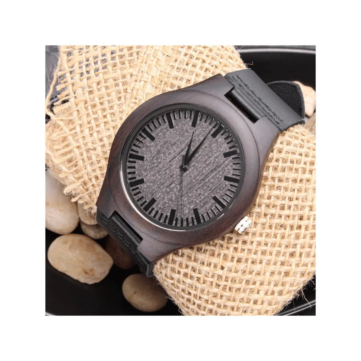 A dark wood watch with a textured gray dial and black strap, displayed on a burlap cloth with small stones.