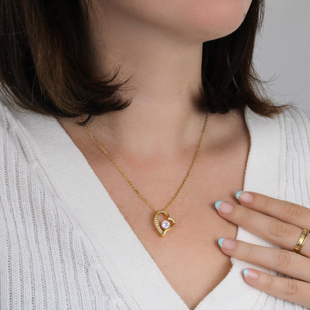 Elegant lifestyle photo of a woman wearing a delicate gold heart-shaped pendant necklace with a sparkling center stone. She is dressed in a cozy white knit sweater, with one hand resting near the necklace. The soft, neutral background and natural styling create a clean, feminine jewelry aesthetic.