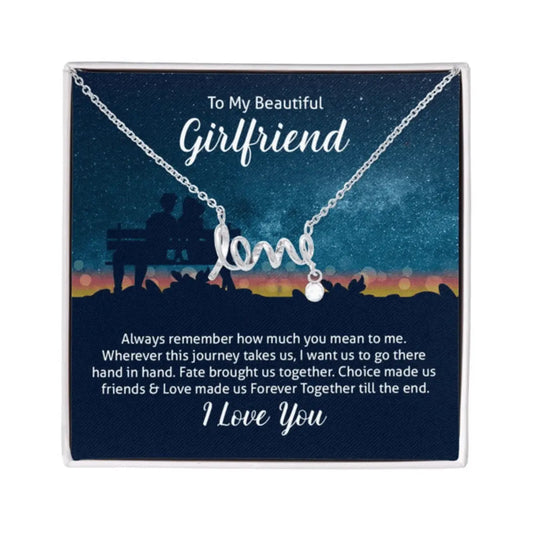 A silver necklace spelling out the word 'love' in cursive, with a small dangling bead, presented on a dark blue gift card with sentimental text for a girlfriend.