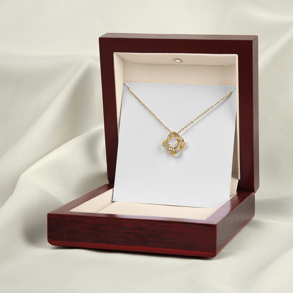 A gold flower-shaped pendant necklace with a clear central gemstone, displayed inside an open dark reddish-brown jewelry box on a cream card against a draped white fabric background.