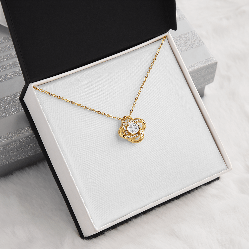 A gold pendant necklace with a clear central gemstone, displayed inside an open black and white jewelry box on a soft white faux-fur surface.