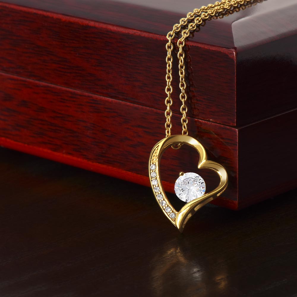 Close-up product photo of a gold heart pendant necklace with an intertwined heart design, sparkling crystal accents, and a large round center stone displayed on a dark red wood box.