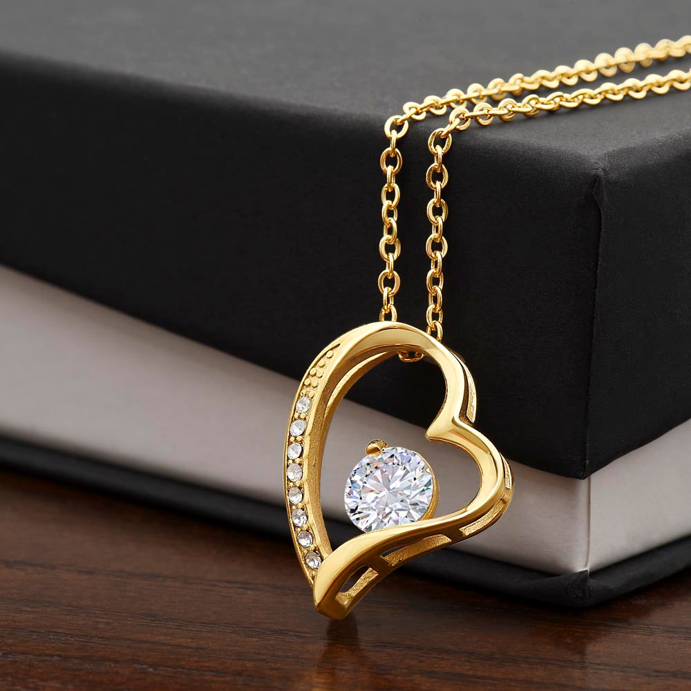 Close-up product photo of a gold heart pendant necklace with an intertwined heart design, sparkling crystal accents, and a large round center stone displayed on a black jewelry box.