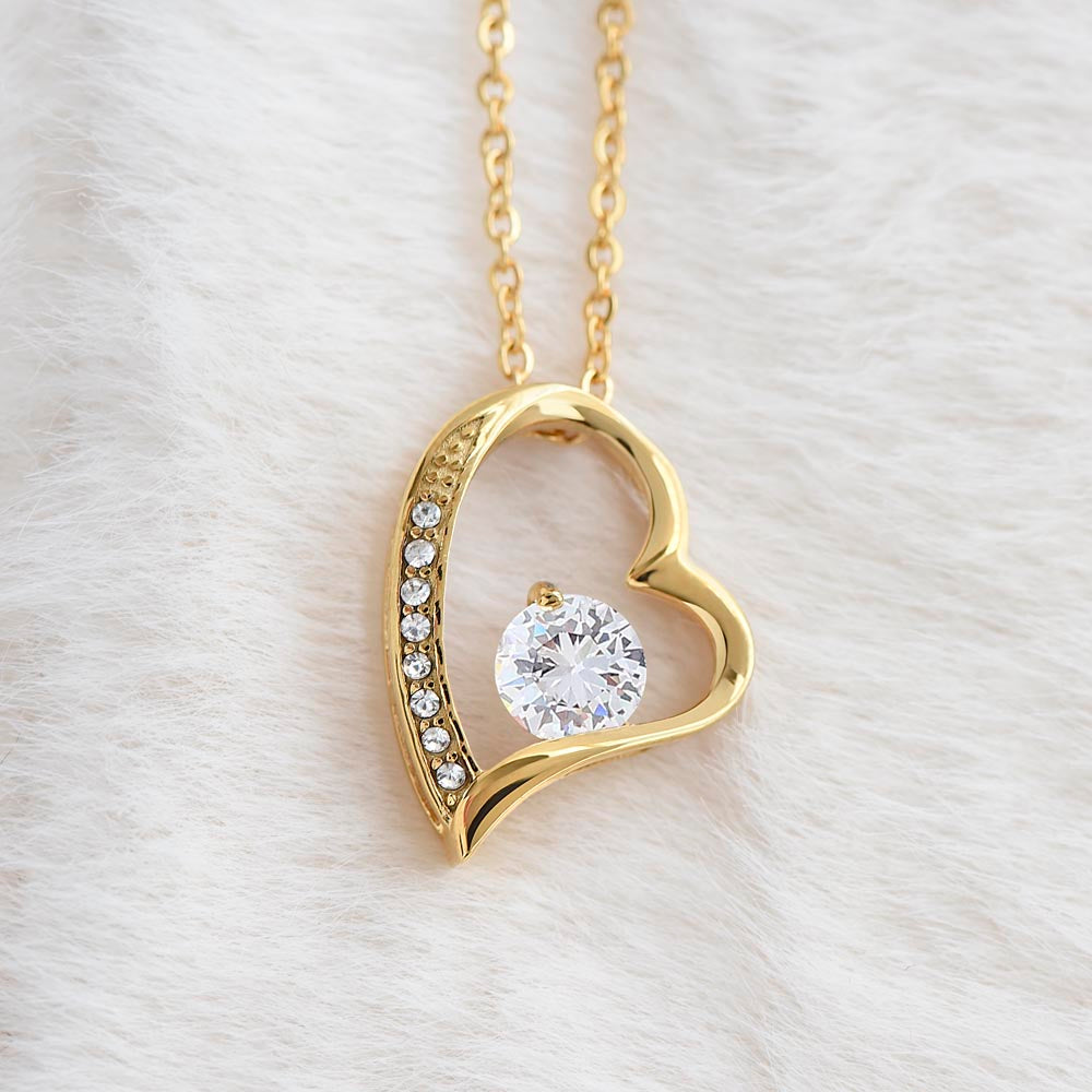 Gold heart-shaped pendant necklace with a large round sparkling gemstone and smaller accent stones, displayed on soft white fur.